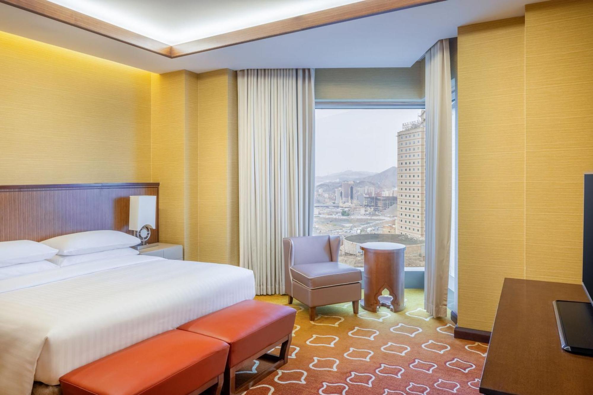 Jabal Omar Marriott Hotel Makkah Mekka Eksteriør billede The photo shows a modern hotel room with a warm color palette. There is a neatly made bed with white sheets and a brown headboard. In front of the bed, there are two red ottomans. A comfortable chair is placed near a window, which offers a view of th