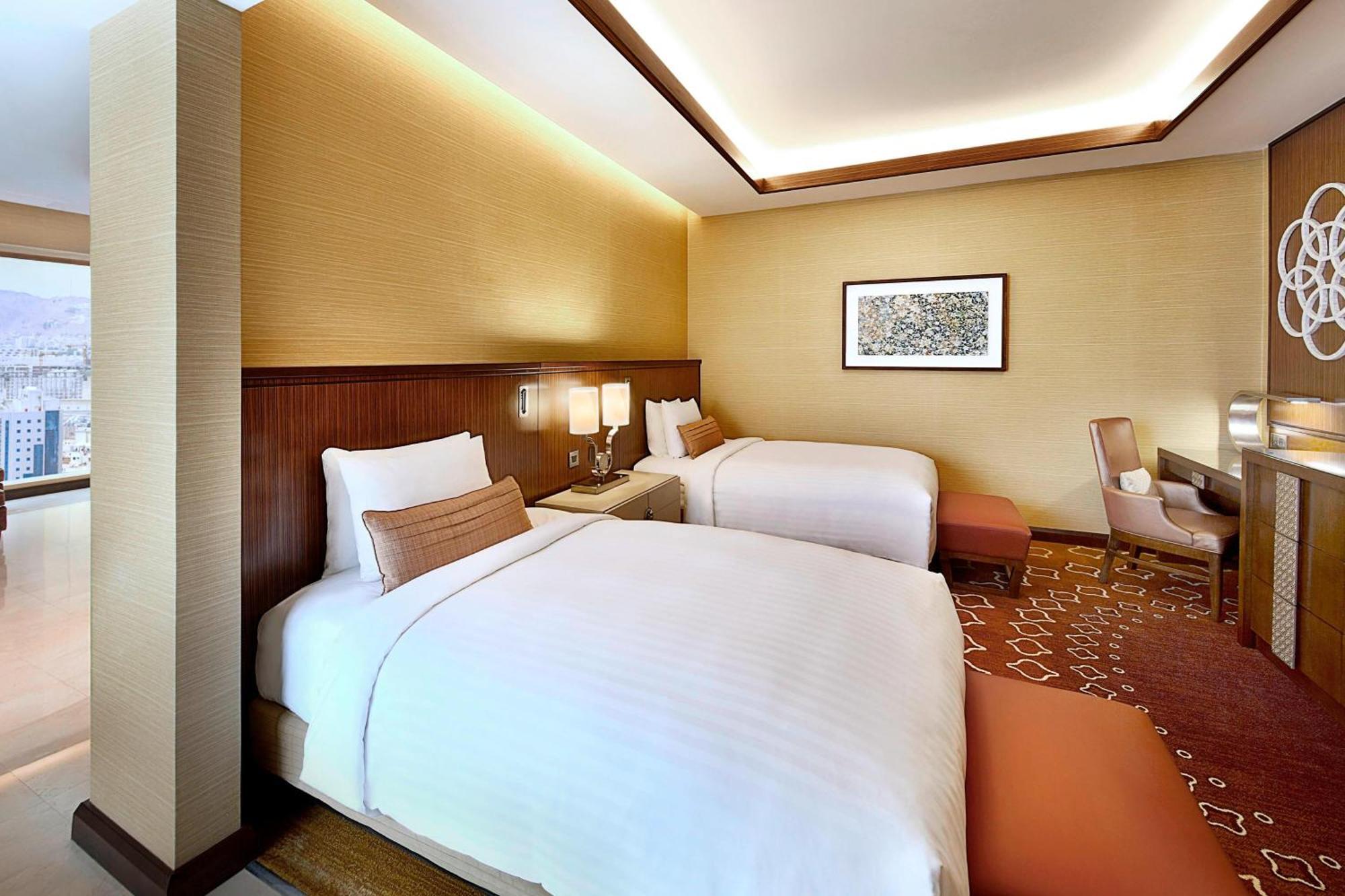 Jabal Omar Marriott Hotel Makkah Mekka Eksteriør billede The photo shows a modern hotel room featuring two beds with white linens. The room has a warm color palette, with beige walls and wooden accents. There's a piece of artwork on the wall above the beds. A small seating area is present, along with a wor
