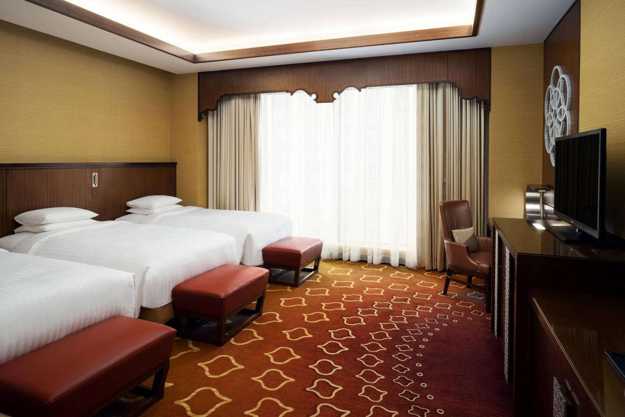 Jabal Omar Marriott Hotel Makkah Mekka Eksteriør billede The photo shows a well-furnished hotel room featuring three beds with white linens. The room has a warm color scheme, with soft yellow walls and a patterned carpet in shades of red and gold. Large windows with light curtains provide natural light, an
